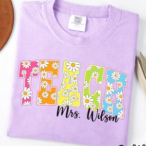 Puede incluir: Una camiseta de color morado claro con la palabra TEACH en letras grandes de bloque rellenas con un patrón de margaritas blancas sobre un fondo colorido. Debajo de la palabra, el texto Mrs. Wilson está impreso en una fuente cursiva negra.