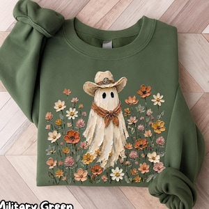 Puede incluir: Una sudadera de cuello redondo en color verde militar con un gráfico central de un fantasma blanco que lleva un sombrero de vaquero beige y un pañuelo marrón. El fantasma está rodeado por un campo de flores pintadas en tonos naranja, rosa y blanco.