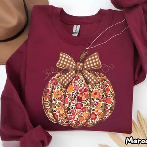 Puede incluir: Una sudadera de cuello redondo y manga larga en color granate con un gráfico central de una calabaza. La calabaza está decorada con un estampado floral colorido en tonos naranja, rojo y amarillo, rematada con un lazo a cuadros marrón y blanco.