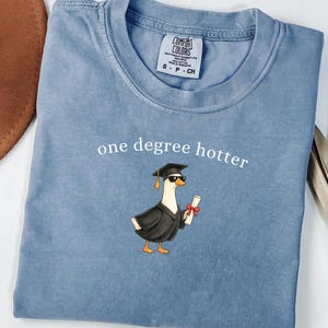 May include: A blue t-shirt featuring a graphic of a duck wearing a black graduation cap and sunglasses while holding a diploma. The text "one degree hotter" is printed in white above the duck. The shirt has a Comfort Colors brand label.