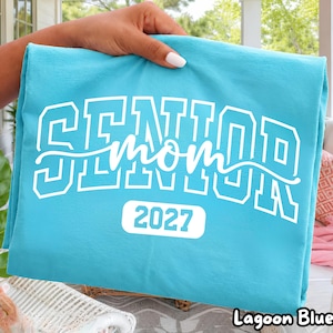 Puede incluir: Una camiseta azul laguna con el texto blanco SENIOR en una fuente universitaria en negrita, con la palabra mom en letra cursiva superpuesta y el número 2027 impreso en un recuadro blanco de esquinas redondeadas en la parte inferior.