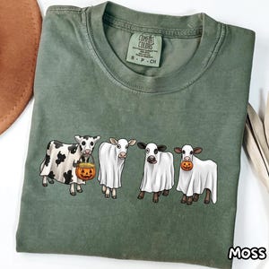 May include: An olive green t-shirt featuring a graphic of four cows wearing white ghost costumes. The cows are standing in a row, with two holding small orange jack-o'-lantern pumpkins. The shirt has a crew neckline and a Comfort Colors brand label.