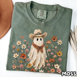 Puede incluir: Una camiseta de cuello redondo color verde salvia con un gráfico de un fantasma con sombrero de vaquero y pañuelo marrón, rodeado de flores naranjas, amarillas y blancas. La etiqueta interior dice Comfort Colors, 100% Cotton, S P CH.