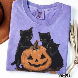 Puede incluir: Una camiseta de manga corta color violeta claro con un gráfico de tres gatitos negros reunidos alrededor de una calabaza naranja tallada. La etiqueta interior dice Comfort Colors, talla S, 100% algodón, hecho en Honduras.