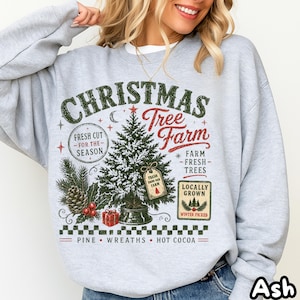 Puede incluir: Una sudadera gris claro de cuello redondo con un estampado gráfico con el texto "Christmas Tree Farm". El diseño incluye un pino central, una piña, una caja de regalo y el texto "Fresh Cut For The Season", "Locally Grown" y "Pine Wreaths Hot Cocoa".