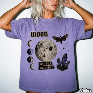 Puede incluir: Una camiseta violeta de manga corta con un estampado gráfico de una luna grande, una polilla y cristales. El texto dice "MOON" y "STAY WILD MOON CHILD" junto a cuatro fases lunares. La camiseta presenta un acabado desteñido de estilo vintage.