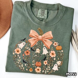 Puede incluir: Una camiseta de cuello redondo color verde musgo con un gráfico en forma de calabaza compuesto por flores, pequeños fantasmas y murciélagos. Un gran lazo color melocotón corona el diseño. La etiqueta interior dice Comfort Colors S P CH.