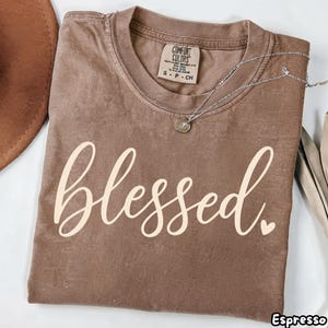 Puede incluir: Una camiseta marrón de manga corta con cuello redondo que presenta la palabra blessed en una tipografía cursiva de color crema claro con un pequeño corazón al final. La etiqueta interior dice Comfort Colors y una fina cadena de plata descansa sobre la tela.