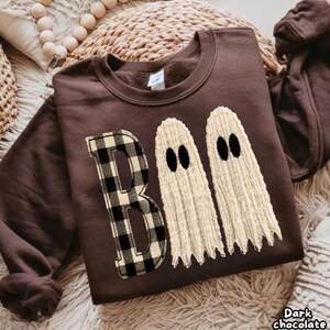 Puede incluir: Una sudadera de cuello redondo en color marrón chocolate oscuro con un diseño gráfico en la parte delantera. El diseño muestra la palabra BOO, donde la letra B tiene un patrón de cuadros blanco y negro, y las dos O son fantasmas color crema texturizados.