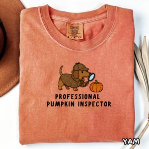 Puede incluir: Una camiseta color coral con el gráfico de un perro salchicha marrón con sombrero y abrigo de detective beige, sosteniendo una lupa junto a una calabaza naranja. El texto debajo dice PROFESSIONAL PUMPKIN INSPECTOR en letras negras.