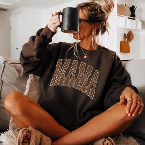 Puede incluir: Una sudadera de manga larga de color marrón oscuro con la palabra MAMA impresa en una fuente grande y arqueada. Las letras son marrones con un patrón de lunares blancos y un borde blanco fino. La sudadera tiene cuello redondo.