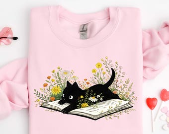 以下が含まれることがあります： 黒猫がカラフルな花に囲まれた開いた本の上に横たわっている、明るいピンク色のスウェットシャツ。猫の体には白いデイジーが描かれています。スウェットシャツは丸いネックラインと長袖です。