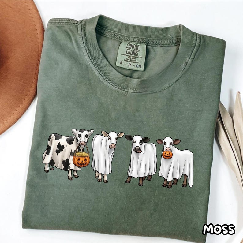 May include: An olive green t-shirt featuring a graphic of four cows wearing white ghost costumes. The cows are standing in a row, with two holding small orange jack-o'-lantern pumpkins. The shirt has a crew neckline and a Comfort Colors brand label.