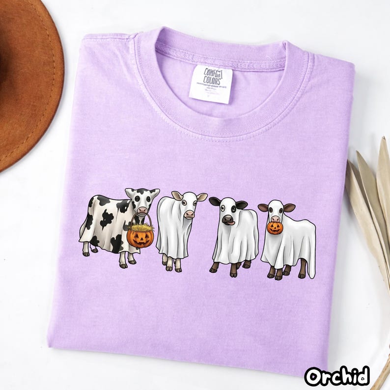 May include: A light purple t-shirt featuring a graphic of four cows wearing white ghost costumes. One cow has black spots, and two of the cows are holding small orange jack-o'-lantern pumpkins. The shirt has a white tag reading Comfort Colors.