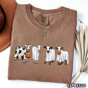 May include: A brown short-sleeve t-shirt featuring a graphic of four cows wearing white ghost costumes. One cow holds a jack-o'-lantern bucket, and another has a jack-o'-lantern in its mouth. The shirt has a Comfort Colors label at the neck.