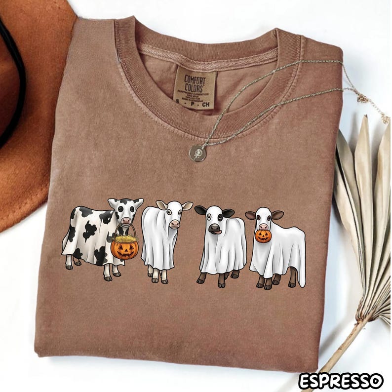 May include: A brown short-sleeve t-shirt featuring a graphic of four cows wearing white ghost costumes. One cow holds a jack-o'-lantern bucket, and another has a jack-o'-lantern in its mouth. The shirt has a Comfort Colors label at the neck.