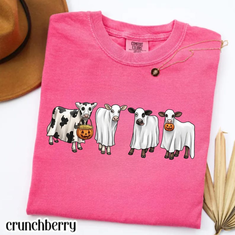 May include: A bright pink short-sleeved t-shirt featuring a graphic of four cows dressed as ghosts. Two of the cows are holding small orange jack-o'-lantern pumpkins. The shirt has a crew neckline and a Comfort Colors brand label at the neck.