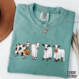 May include: A light green short-sleeve t-shirt featuring a graphic of four cows wearing white ghost costumes. One cow holds a jack-o'-lantern bucket. The shirt has a crew neckline and a Comfort Colors brand label printed on the inside collar.
