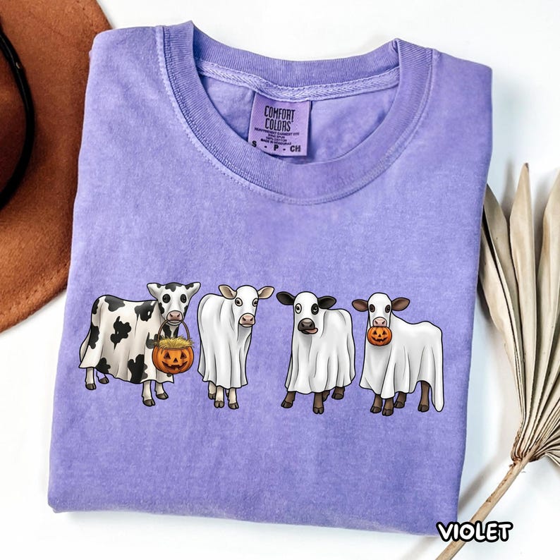 May include: A violet short-sleeved t-shirt featuring a graphic of four cows wearing white ghost costumes. One cow holds a pumpkin bucket filled with hay, and another holds a small pumpkin in its mouth. The shirt has a Comfort Colors brand label.