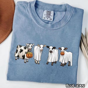 May include: A blue short-sleeve t-shirt featuring a graphic of four cows wearing white ghost costumes. One cow holds a jack-o'-lantern bucket filled with hay, and another holds a small pumpkin in its mouth. The shirt has a Comfort Colors brand label.