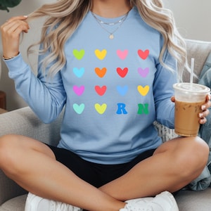 May include: A light blue long-sleeve shirt with a pattern of colorful hearts. The shirt also features the letters "R N" in green and blue. The shirt is made of a soft material.