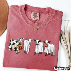 May include: A faded crimson short-sleeve t-shirt featuring a graphic of four cows dressed as ghosts. The cows are depicted wearing white sheets, with one holding a jack-o'-lantern bucket and another holding a small pumpkin. The shirt has a crew neckline.