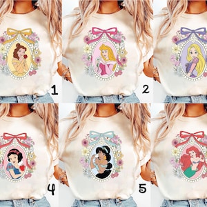 May include: Cream-colored t-shirts featuring six different princess designs. Each design is framed by a floral oval and a ribbon. The princesses include Belle, Aurora, Rapunzel, Snow White, Jasmine, and Ariel.