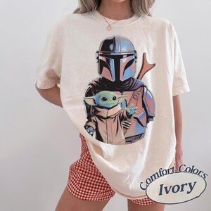 May include: An ivory short-sleeve t-shirt featuring a graphic print of a silver-helmeted armored character standing behind a small green creature with large ears and a tan robe. The shirt has a crew neckline and a relaxed, loose-fitting silhouette.