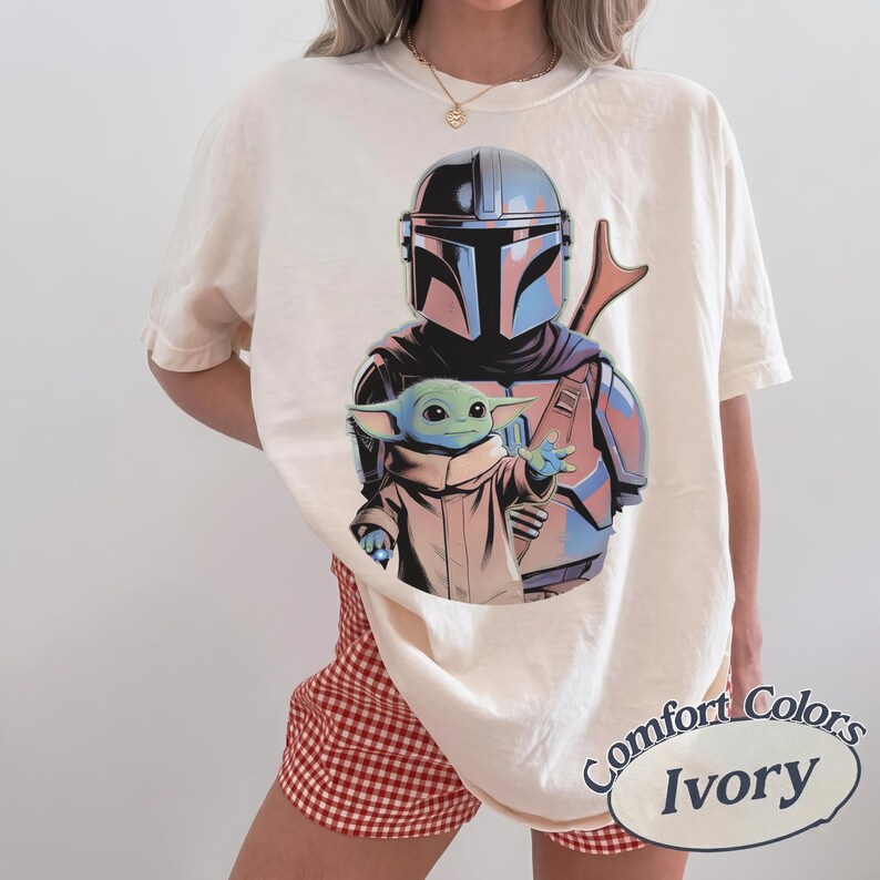 May include: An ivory short-sleeve t-shirt featuring a graphic print of a silver-helmeted armored character standing behind a small green creature with large ears and a tan robe. The shirt has a crew neckline and a relaxed, loose-fitting silhouette.
