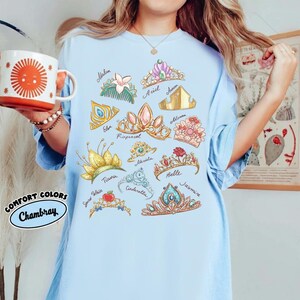 May include: Light blue t-shirt featuring a collection of illustrated princess tiaras with names like Ariel, Belle, and Jasmine. The shirt also has the words "Comfort Colors Chambray" in a circular design.
