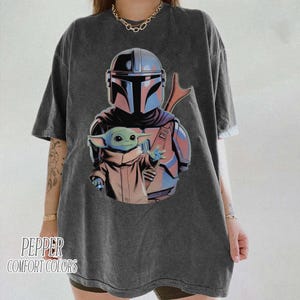 May include: A charcoal gray short-sleeve t-shirt featuring a graphic print of a helmeted warrior in blue and silver armor holding a small green creature in a tan robe. The text PEPPER COMFORT COLORS is printed in the bottom left corner of the shirt.