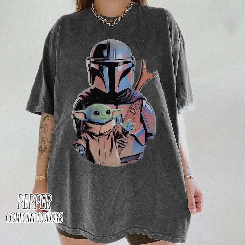 May include: A charcoal gray short-sleeve t-shirt featuring a graphic print of a helmeted warrior in blue and silver armor holding a small green creature in a tan robe. The text PEPPER COMFORT COLORS is printed in the bottom left corner of the shirt.
