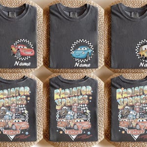 May include: Six dark gray t-shirts with different Cars movie graphics. Three shirts feature a car character with the word "Name" below. The other three shirts have a "Radiator Springs" design with various Cars characters.