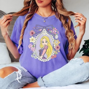 May include: A lilac-colored t-shirt featuring a framed illustration of a cartoon princess with long blonde hair. The frame is decorated with flowers and a bow. The princess is wearing a purple dress with pink sleeves.