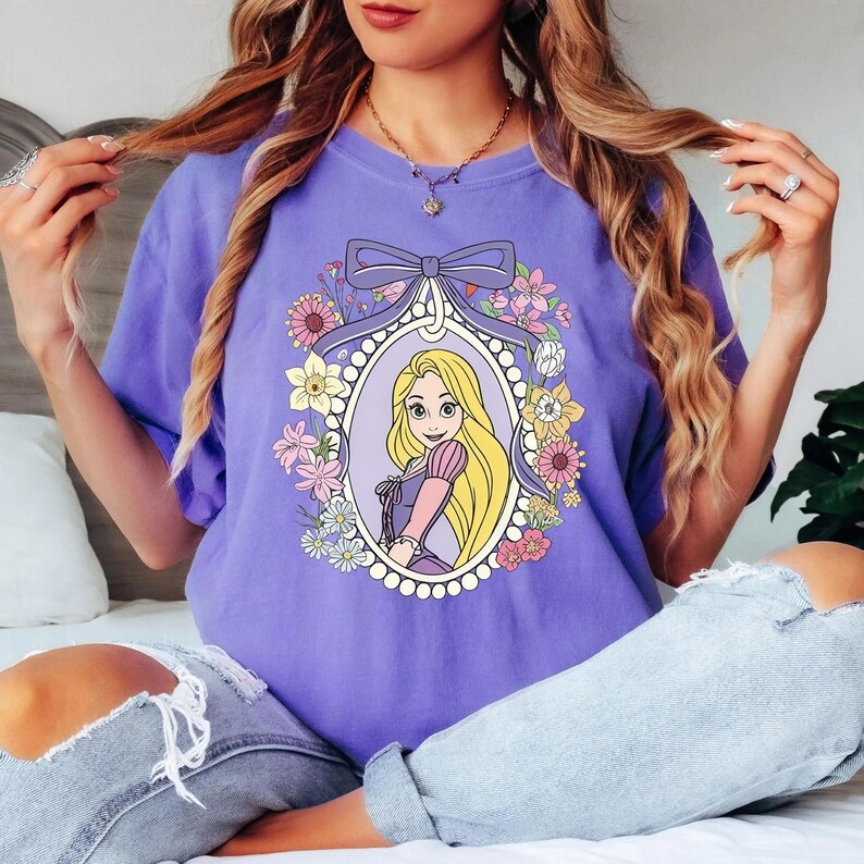 May include: A lilac-colored t-shirt featuring a framed illustration of a cartoon princess with long blonde hair. The frame is decorated with flowers and a bow. The princess is wearing a purple dress with pink sleeves.