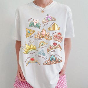May include: White t-shirt featuring colorful illustrations of various princess tiaras, each labeled with a name. The designs include crowns for Ariel, Aurora, Belle, Cinderella, Elsa, Jasmine, Mulan, Moana, Rapunzel, Snow White, and Tiana.