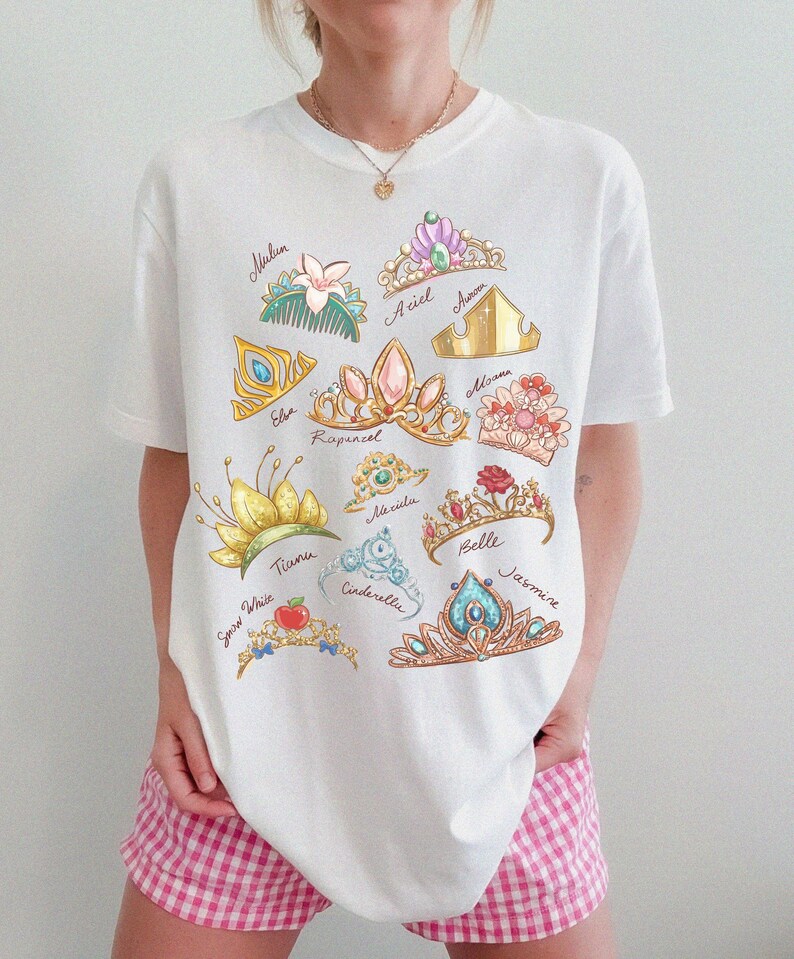 May include: White t-shirt featuring colorful illustrations of various princess tiaras, each labeled with a name. The designs include crowns for Ariel, Aurora, Belle, Cinderella, Elsa, Jasmine, Mulan, Moana, Rapunzel, Snow White, and Tiana.