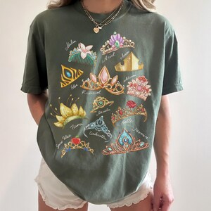 May include: Olive green t-shirt featuring colorful illustrations of various princess tiaras, each labeled with a princess's name. The tiaras are in gold, pink, blue, and other colors. The shirt is a casual, comfortable style.
