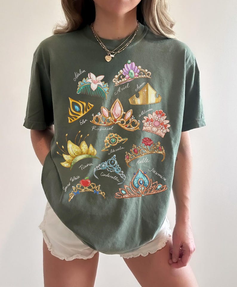 May include: Olive green t-shirt featuring colorful illustrations of various princess tiaras, each labeled with a princess's name. The tiaras are in gold, pink, blue, and other colors. The shirt is a casual, comfortable style.