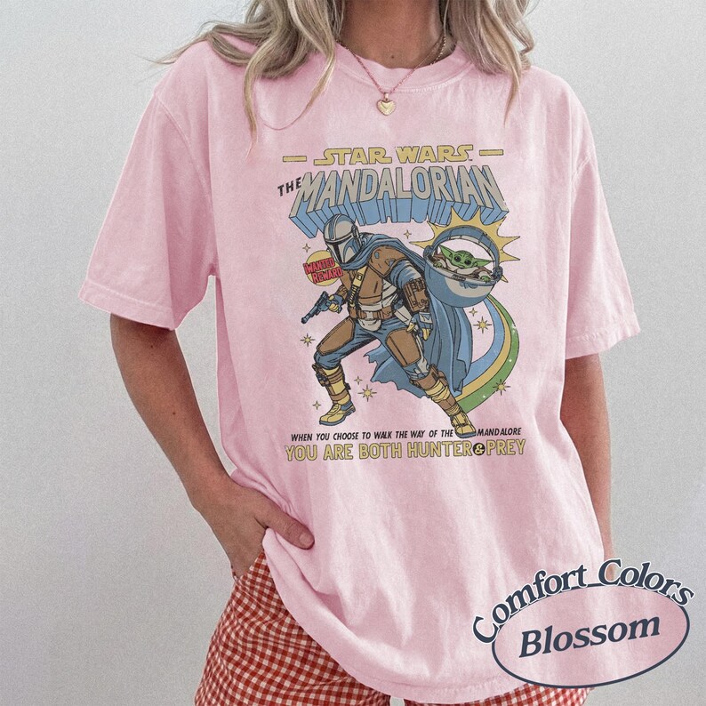 May include: Pink t-shirt featuring a graphic from "The Mandalorian" with the text "STAR WARS THE MANDALORIAN" and the quote "WHEN YOU CHOOSE TO WALK THE WAY OF THE MANDALORE YOU ARE BOTH HUNTER PREY". The shirt has a Comfort Colors Blossom label.