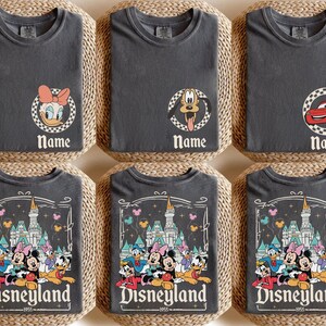 May include: Six grey t-shirts featuring Disney character designs. Three shirts have character portraits with the word "Name" below. The other three shirts feature a Disneyland graphic with the text "Disneyland 1955".
