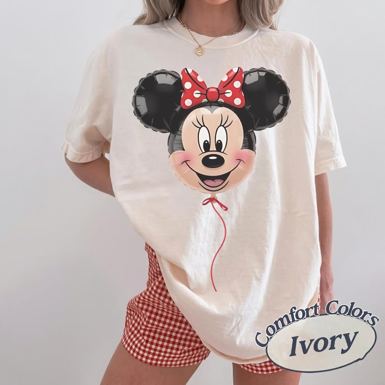 May include: Ivory-colored t-shirt featuring a Minnie Mouse balloon design. The balloon has a red bow with white polka dots and a red string. The shirt is a Comfort Colors brand.