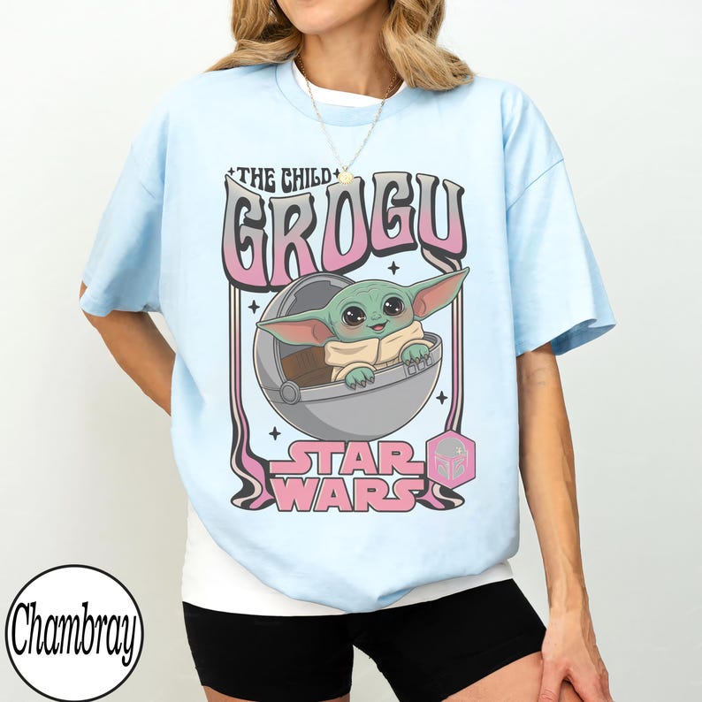 Puede incluir: Una camiseta azul claro con un gr&aacute;fico de Grogu de Star Wars sentado en una c&aacute;psula flotante. El texto dice THE CHILD GROGU y STAR WARS en letras rosas y negras. El dise&ntilde;o incluye peque&ntilde;os detalles de estrellas y un icono hexagonal rosa.