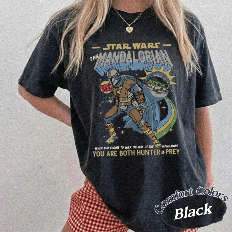 May include: Black t-shirt featuring a graphic of The Mandalorian and Grogu from Star Wars. The design includes text that reads "The Mandalorian" and "You are both hunter & prey." The shirt is labeled "Comfort Colors Black."