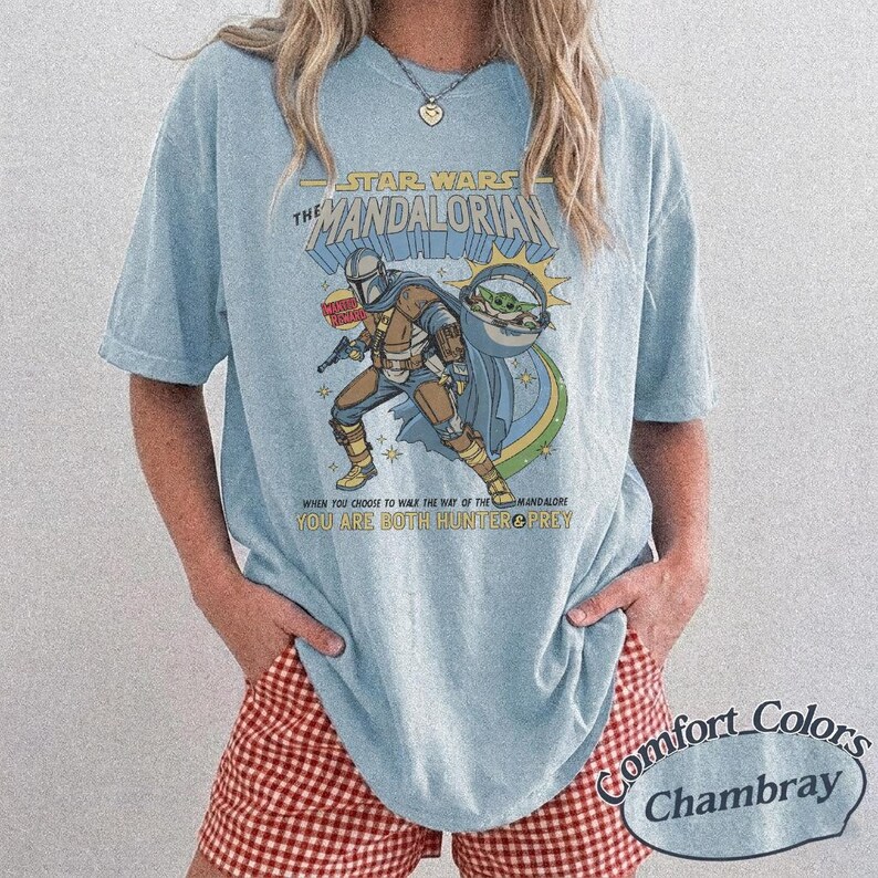 May include: Light blue t-shirt featuring a Star Wars Mandalorian graphic. The design includes the Mandalorian, Grogu, and text that reads "When you choose to walk the way of the Mandalorian, you are both hunter & prey."