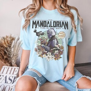 May include: Light blue t-shirt featuring a graphic of The Mandalorian and Grogu surrounded by flowers. The text "THE MANDALORIAN THIS IS THE WAY" is printed across the top of the design. The shirt is made of a soft material.