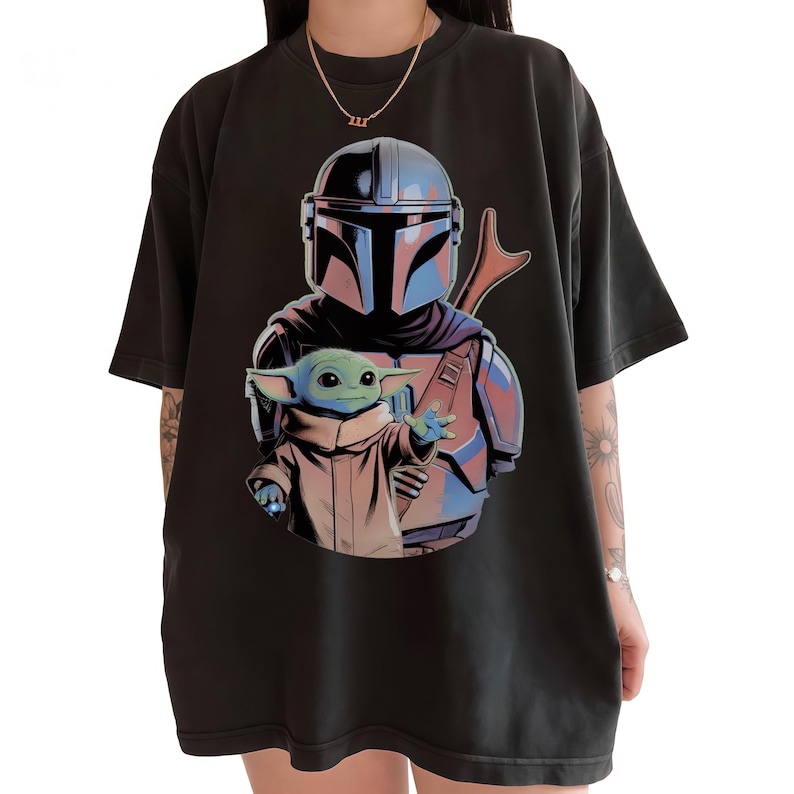 May include: A black short-sleeved t-shirt featuring a graphic print of a helmeted character in silver and blue armor standing behind a small green creature with large ears wearing a tan robe, with a brown rifle visible over the character's shoulder.