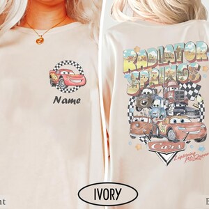 May include: Ivory-colored long-sleeve shirt featuring a cartoon car design on the front and back. The front displays a red car within a checkered circle, with the word "Name" below. The back showcases "Radiator Springs" and other car characters.