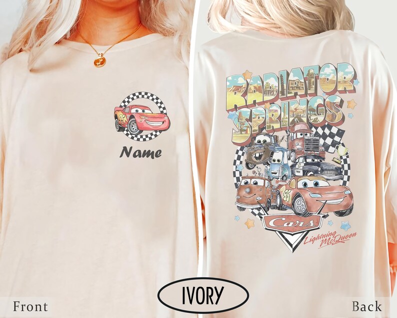 May include: Ivory-colored long-sleeve shirt featuring a cartoon car design on the front and back. The front displays a red car within a checkered circle, with the word "Name" below. The back showcases "Radiator Springs" and other car characters.
