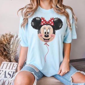 May include: Light blue t-shirt featuring a Minnie Mouse balloon graphic. The balloon has black ears, a red bow with white polka dots, and a smiling face. A red string hangs from the balloon. The shirt is a light blue color.